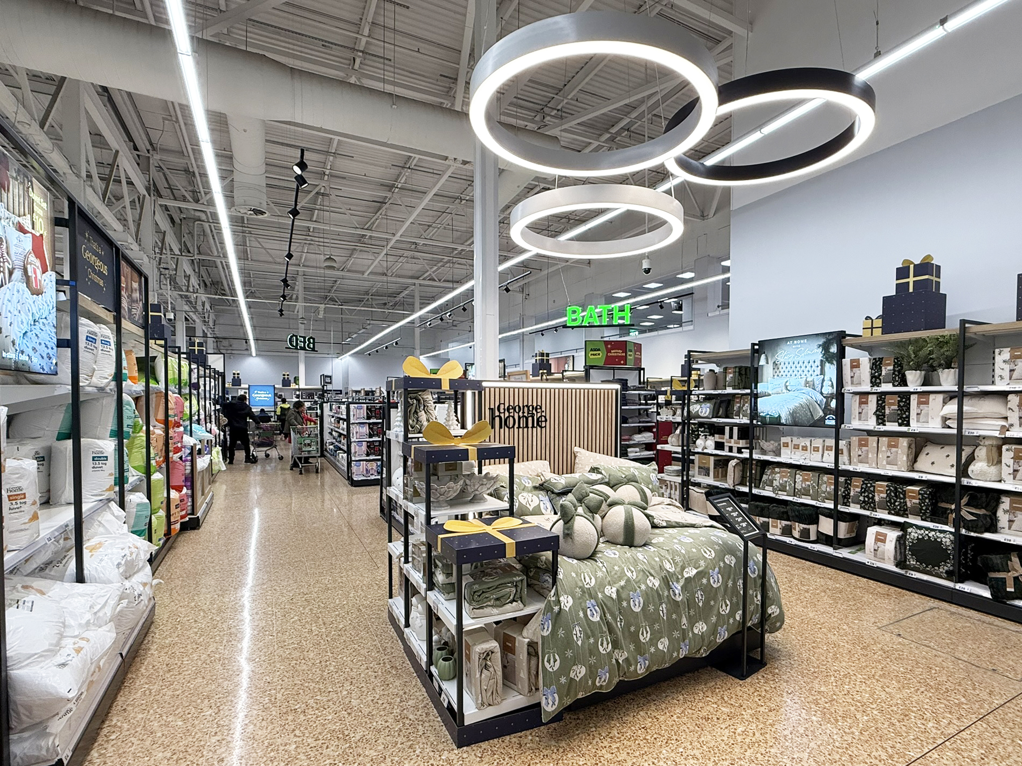 Aisle in a home goods store with bedding on shelves and a bed display in the foreground and ceiling ring lights overhead