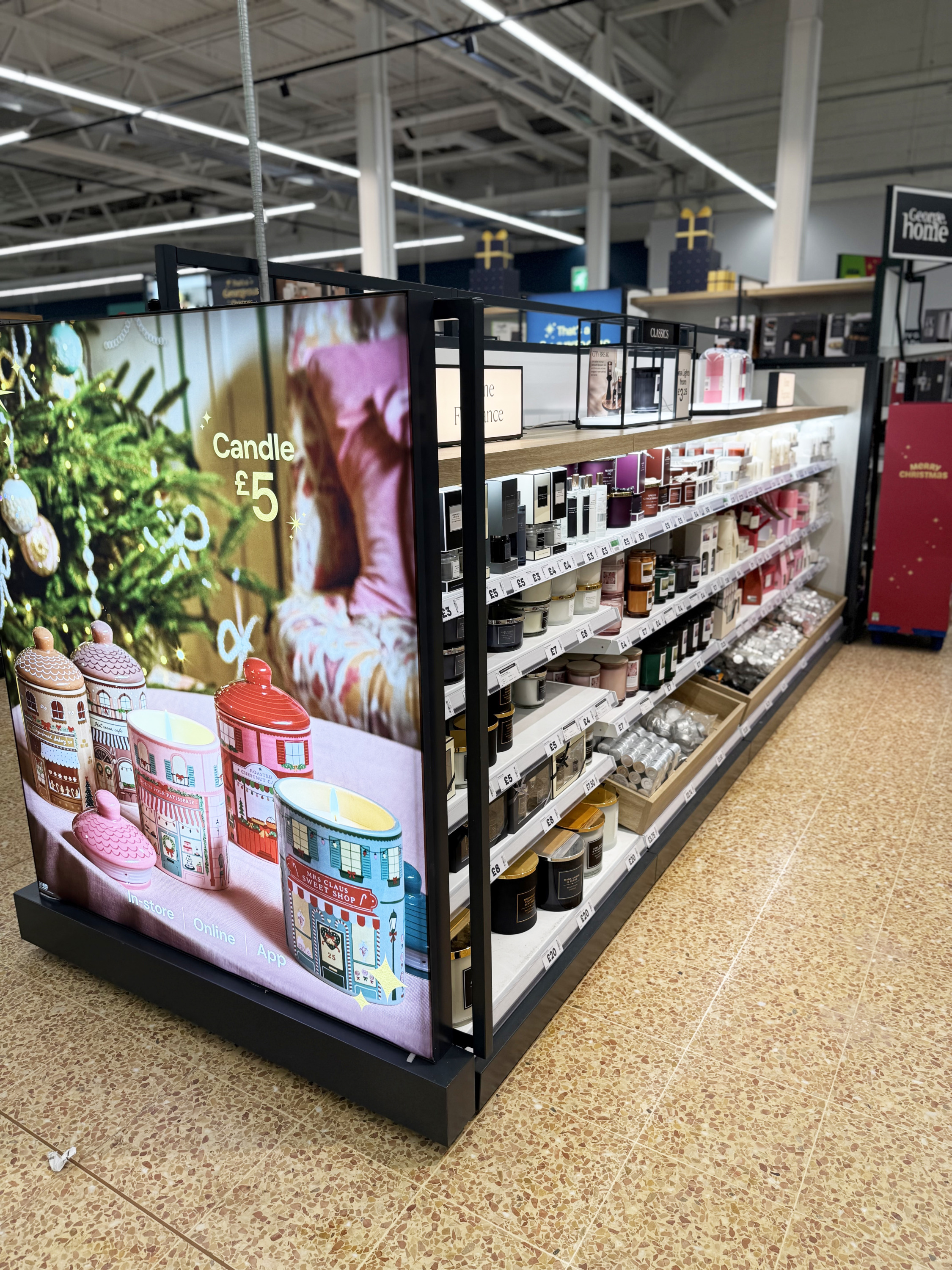 Store aisle with shelves of candles and cosmetics; festive display panel with teapots and a Christmas scene at the front.